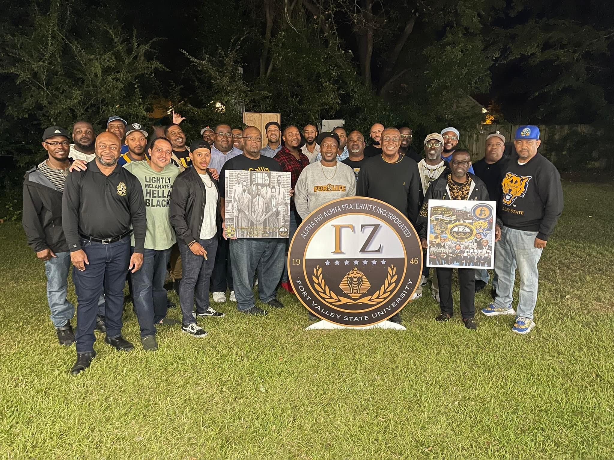 Brothers gathered at night around the Gamma Zeta chapter seal with framed chapter history.