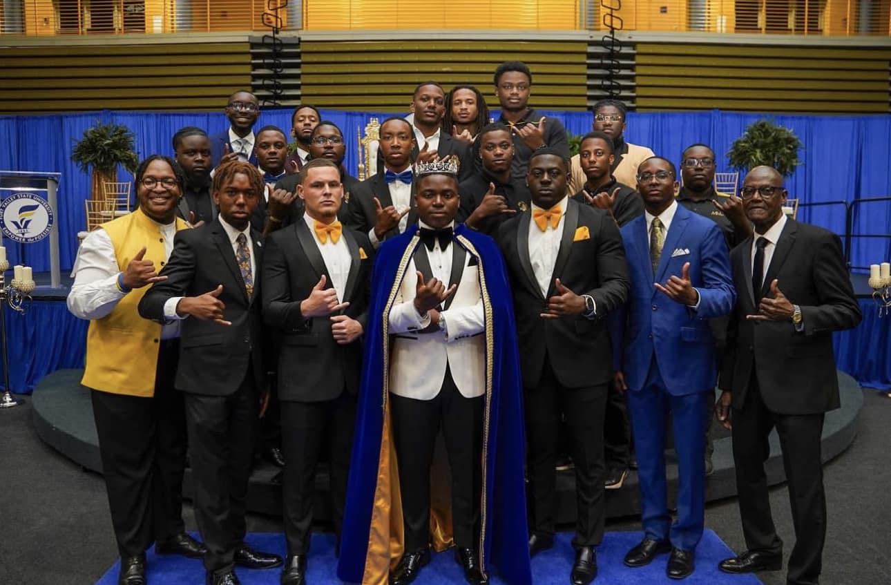 Gamma Zeta brothers in formal attire surrounding the crowned homecoming king in his royal blue cape.