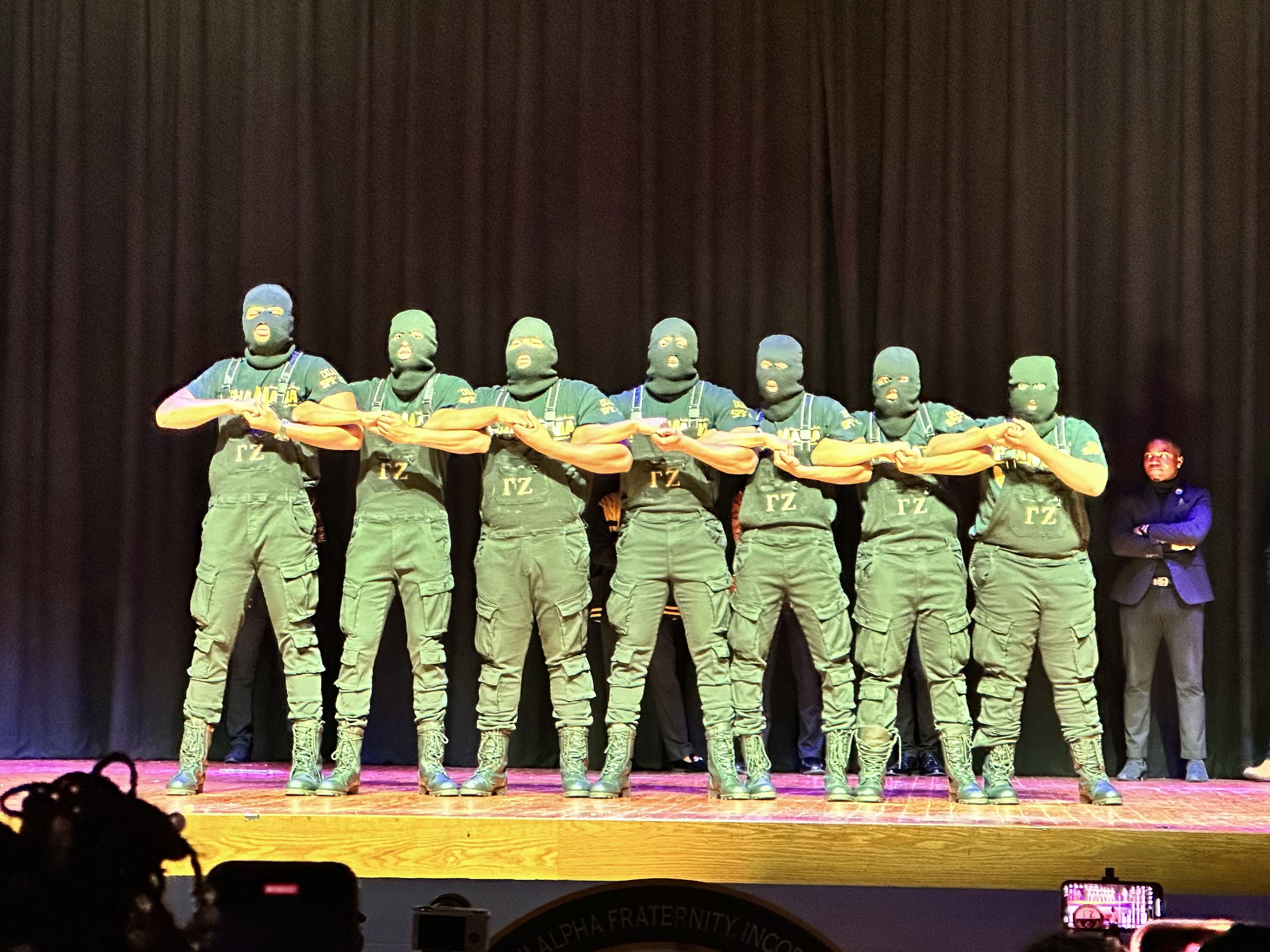 The Spring 2023 Alphamania line under green stage lighting, seven brothers in green ski masks, overalls and cargo pants marked with the Gamma Zeta letters, linked arm-in-arm in a step formation.