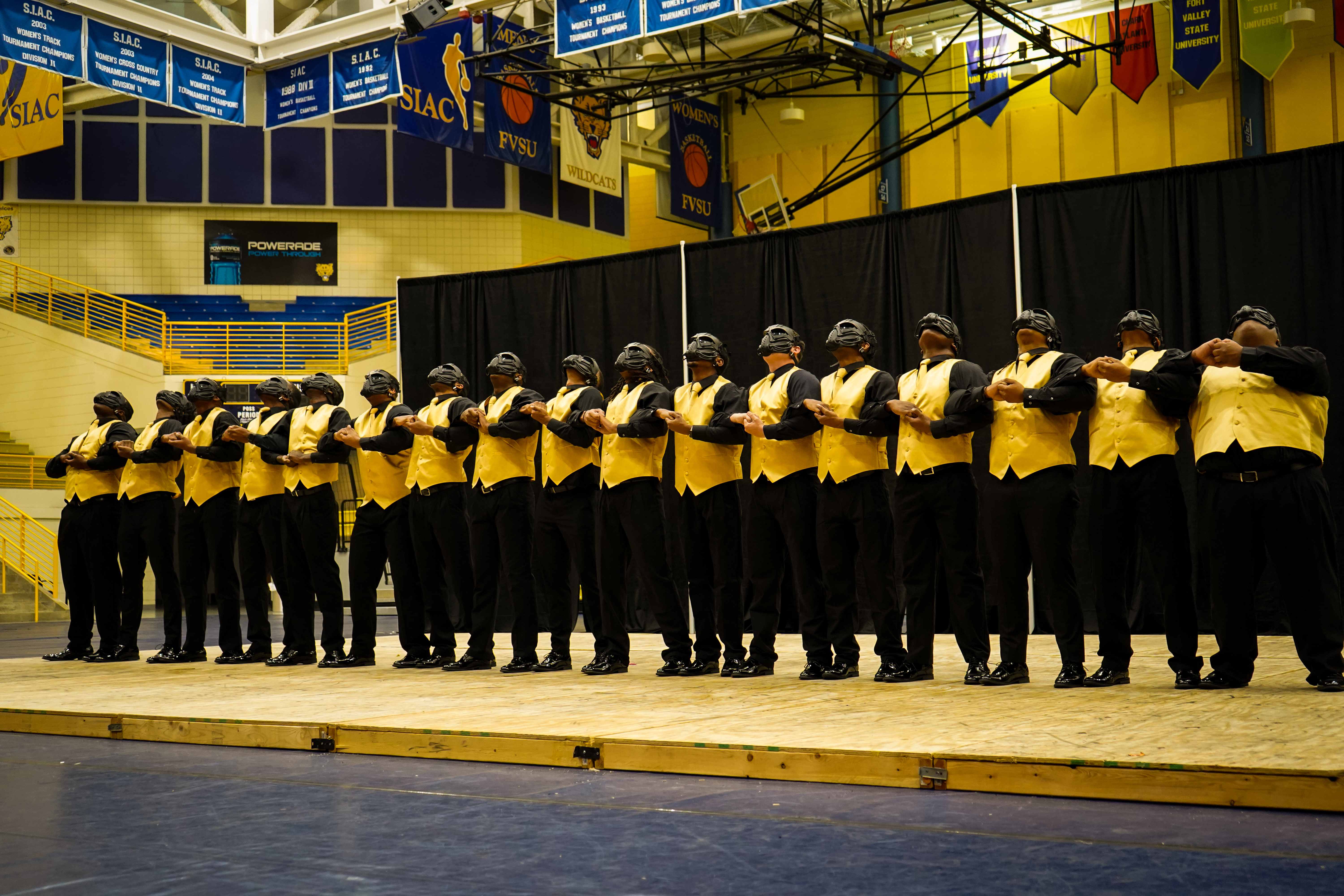 The Spring 2022 Galactic I.C.E. line in a long arm-linked step formation on a gym stage, wearing gold vests over black shirts and masks, with FVSU and SIAC championship banners overhead.
