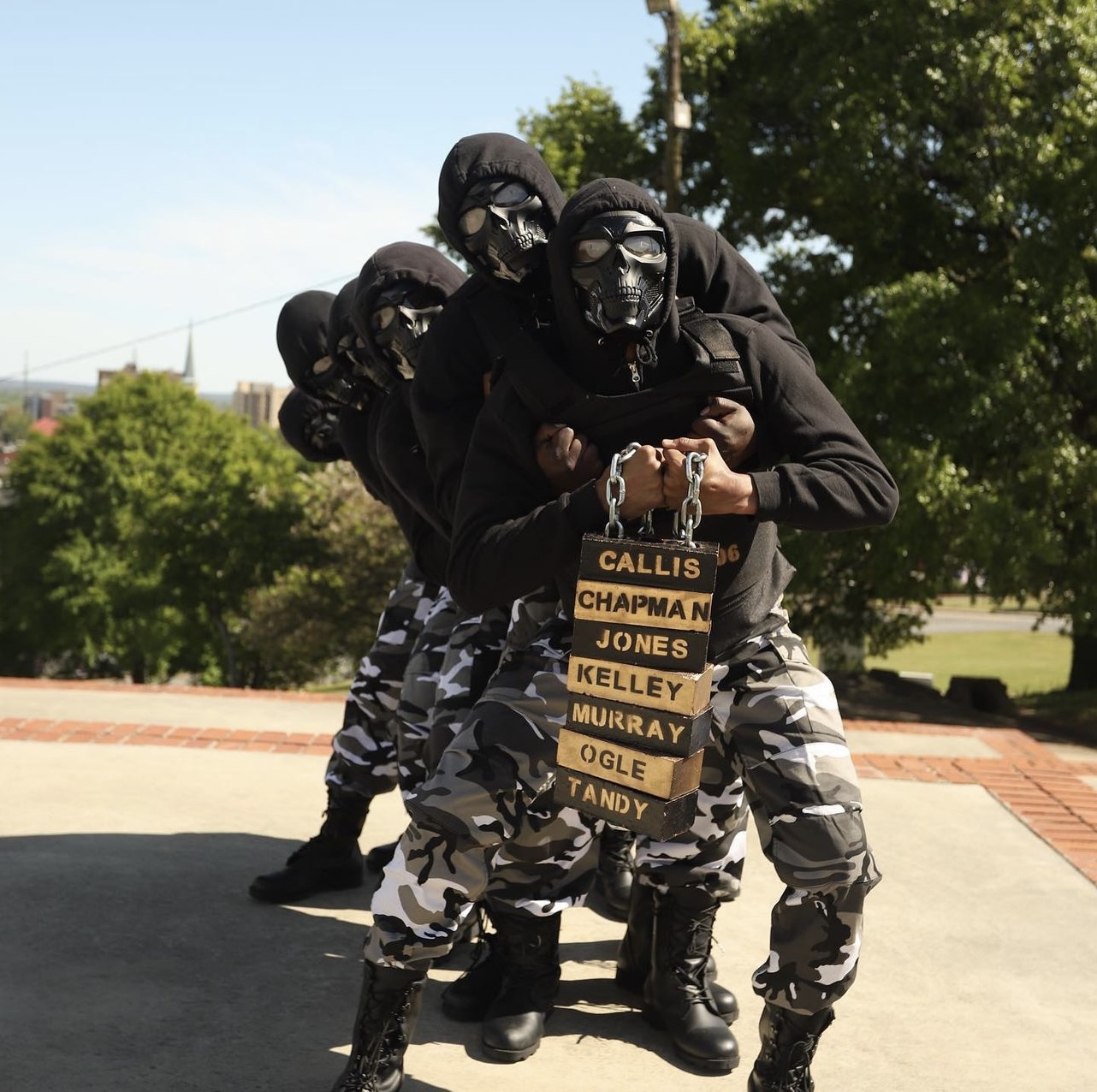 The Spring 2021 H.E.I.R.S. line in step formation, dressed in black hoodies and skull masks with camo pants and combat boots, holding a chained stack of wooden name-plaques reading Callis, Chapman, Jones, Kelley, Murray, Ogle, and Tandy.