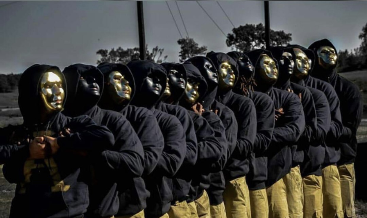 The Spring 2016 K.R.I.T.T line posed outdoors in a diagonal row, wearing black hoodies, khaki pants, and shiny gold face masks with arms folded across their chests.