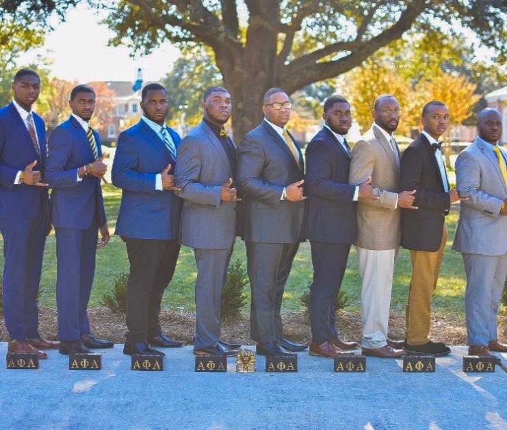 The Fall 2019 Mission: Possible line posed in a diagonal row on a campus lawn, dressed in suits, with black Alpha Phi Alpha name-blocks set on the ground at their feet.