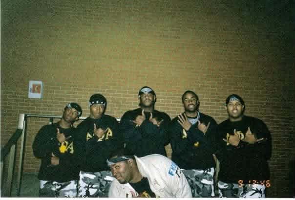 The Fall 2000 New I.C.E. Age line posed against a brick wall in black Alpha Phi Alpha sweatshirts and gray camo pants throwing hand signs, with one brother crouched in front.