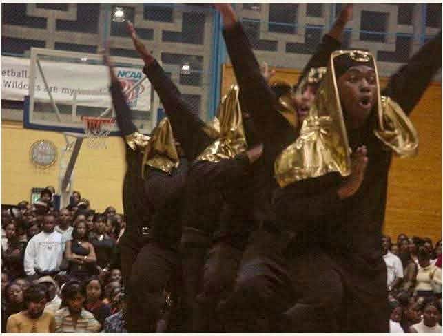 The Fall 2000 New I.C.E. Age line performing at a step show in a packed gym, dressed in all black with gold hoods, arms thrown up mid-step beneath a basketball hoop.