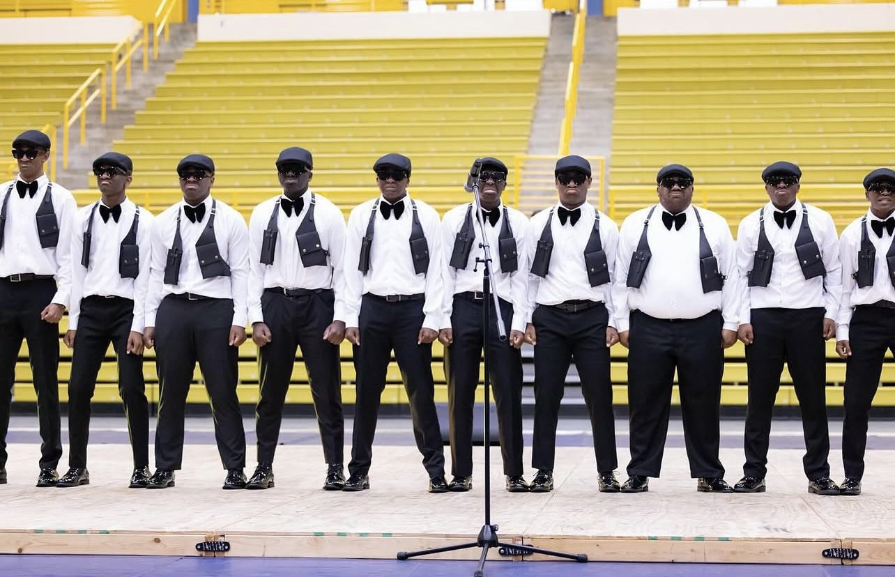 The Spring 2024 N.O.B.L.E. Narratives line on a probate stage in an arena with yellow bleachers, standing in a row in matching white shirts, black bow ties, tactical vests, newsboy caps, and sunglasses behind a microphone stand.