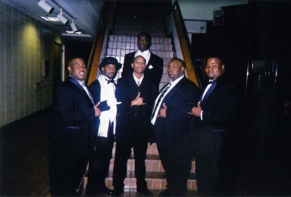 The Fall 1999 Phi 2 K line in tuxedos and suits posed on a staircase in a dim hallway, giving Alpha hand signs.