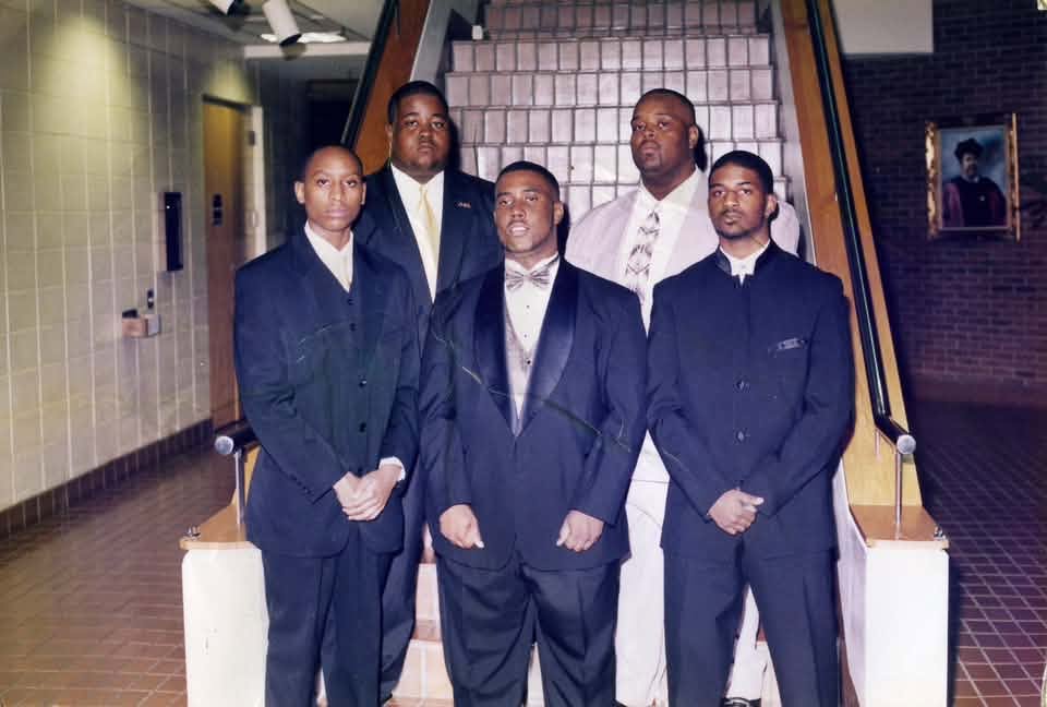 The Fall 1999 Phi 2 K line in formal suits and tuxedos posed at the foot of a staircase in a brick building.