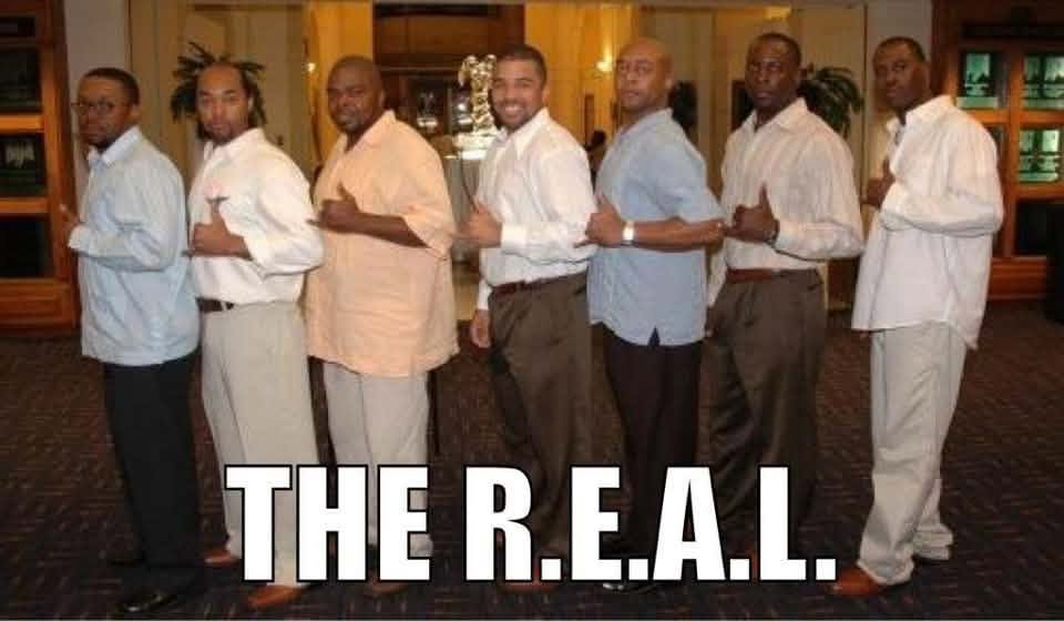 The Fall 1995 Revival of the R.E.A.L. line standing in a diagonal row inside a hotel hall, dressed in dress shirts and slacks, each giving the Alpha hand sign.