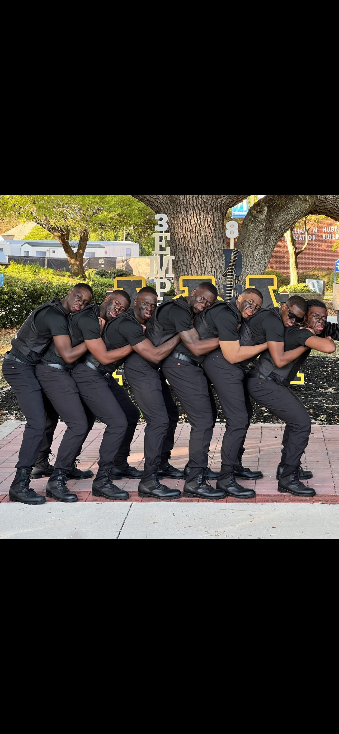 The Spring 2025 VII S.A.L.V.O. line in a tight single-file step formation, dressed in black shirts, tactical pants, and combat boots, posed under a large campus tree with black-and-gold Greek-letter stands behind them.