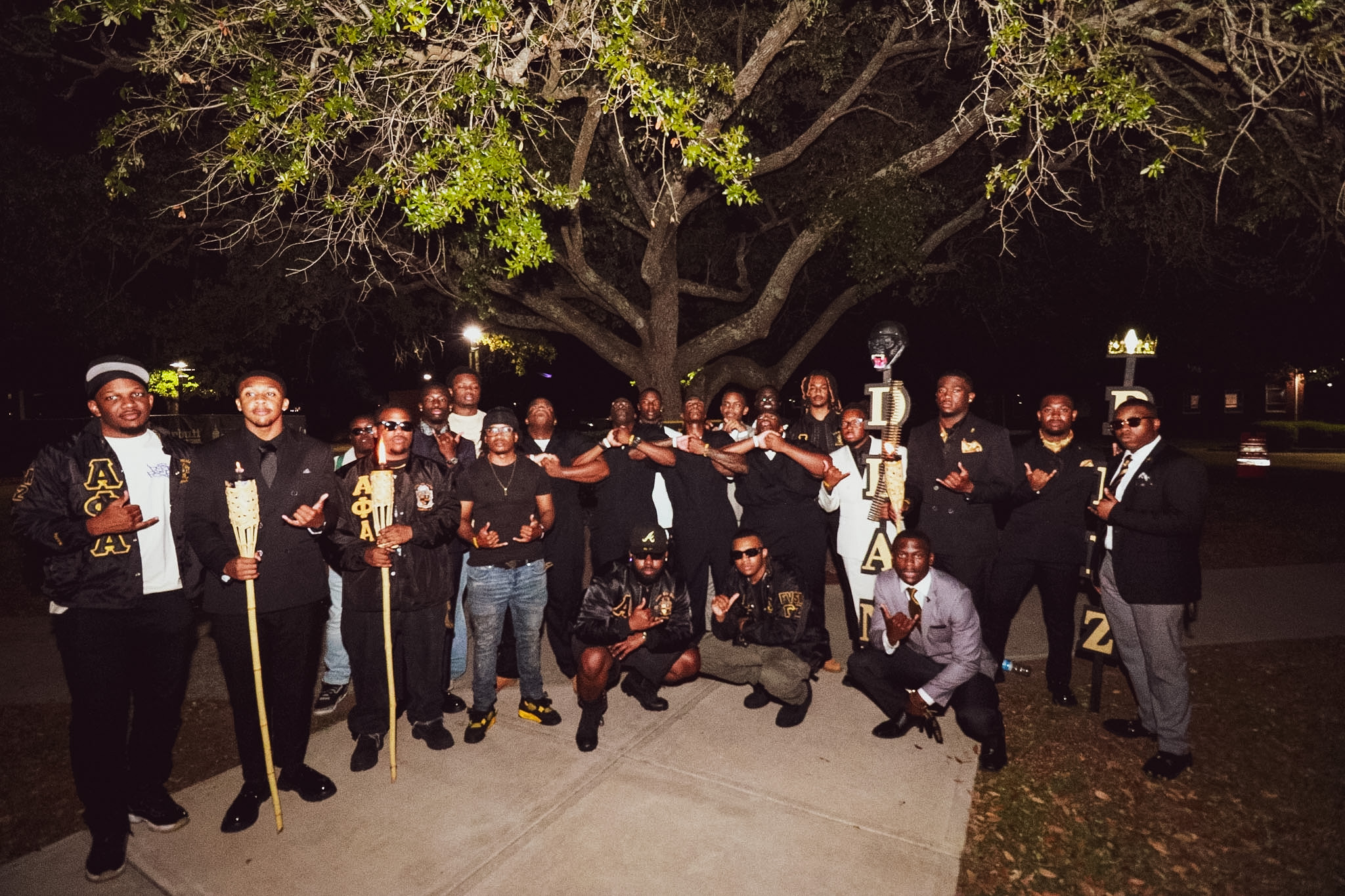 The Spring 2026 line and Gamma Zeta brothers gathered at night under a large oak tree on campus, dressed in Alpha Phi Alpha black and gold with lit bamboo torches, throwing the fraternity hand sign.