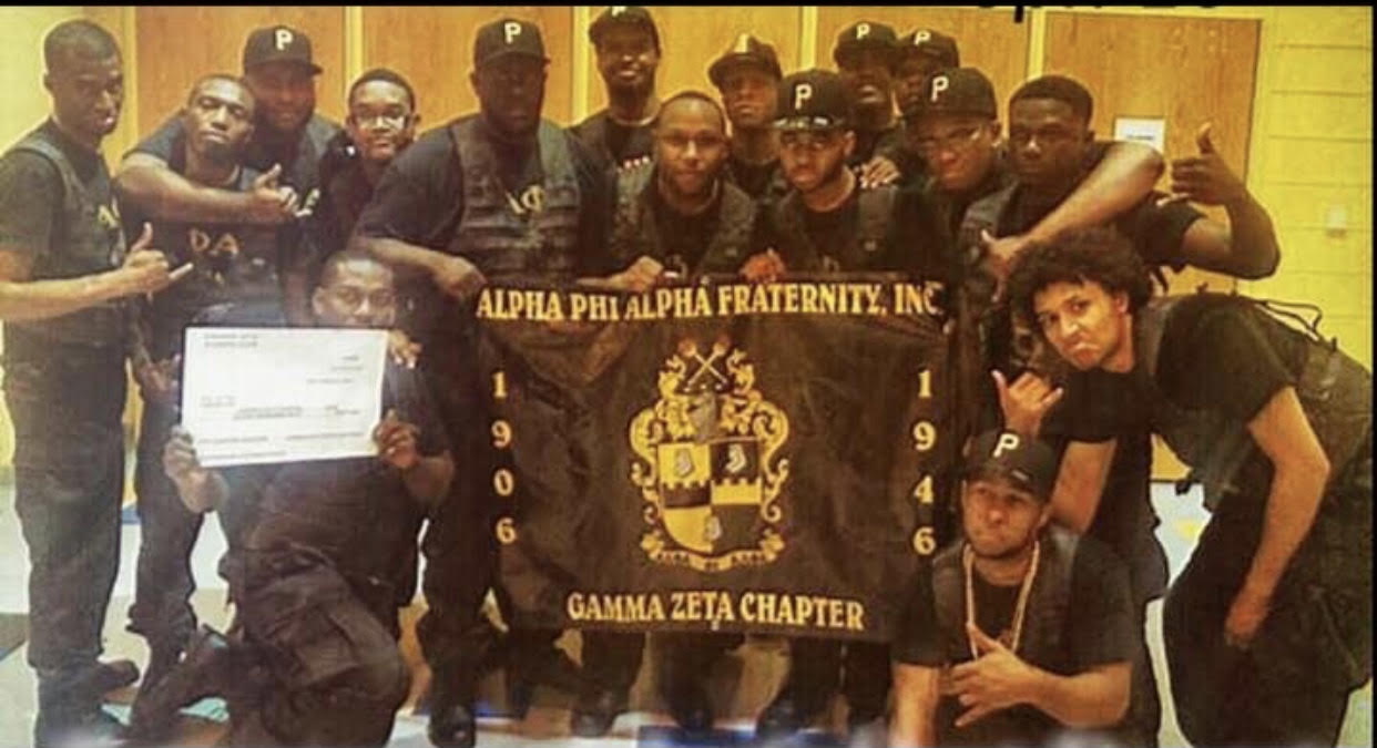 The Spring 2015 T.R.O.T.C.D line and Gamma Zeta brothers gathered in a gym around a large black-and-gold Alpha Phi Alpha Fraternity, Inc. Gamma Zeta Chapter banner, dressed in black step attire and P caps, with one member holding a certificate.